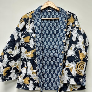 May include: A quilted jacket with a navy blue exterior featuring white and gold crane illustrations. The interior lining is a blue paisley pattern. The jacket has a button-up front and is hanging on a wooden hanger.