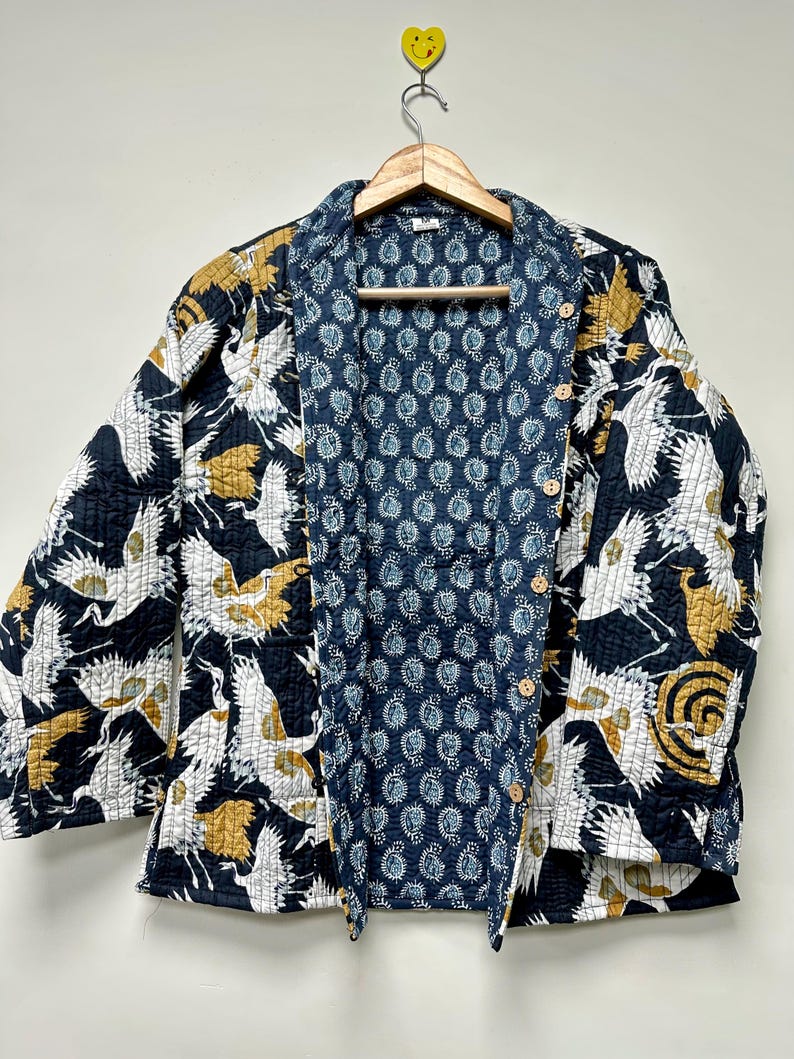 May include: A quilted jacket with a navy blue exterior featuring white and gold crane illustrations. The interior lining is a blue paisley pattern. The jacket has a button-up front and is hanging on a wooden hanger.