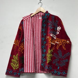 Könnte beinhalten: Eine dunkelrote Samtjacke mit farbenfrohen Stickereien von Leoparden und Blattwerk. Die Jacke hat einen Stehkragen und eine offene Vorderseite, die ein rot-weiß gestreiftes Innenfutter zeigt. Die Ärmel und Vorderteile sind aufwendig verziert.