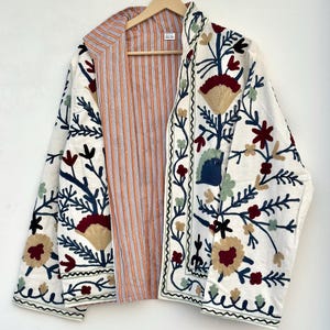 May include: A long-sleeved jacket featuring an off-white base with intricate floral embroidery in shades of dark red, navy blue, and sage green. The interior lining displays a vertical orange and white striped pattern with a small white label.
