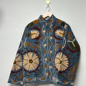 May include: A blue velvet jacket featuring intricate floral embroidery in shades of cream, dark red, and olive green. The jacket has a high collar, long sleeves, and a front opening, showcasing detailed botanical patterns across the entire surface.