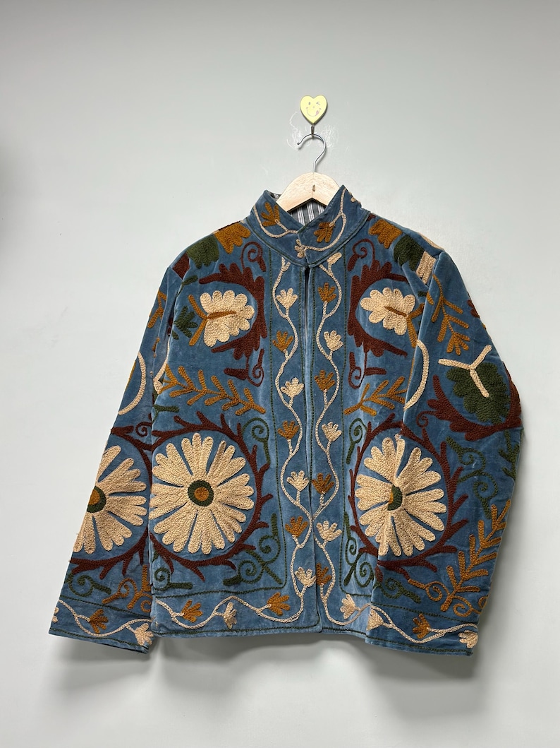 May include: A blue velvet jacket featuring intricate floral embroidery in shades of cream, dark red, and olive green. The jacket has a high collar, long sleeves, and a front opening, showcasing detailed botanical patterns across the entire surface.