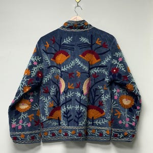 May include: A blue jacket featuring intricate, multi-colored floral embroidery. The design includes large orange, pink, and dark red flower motifs with white leafy stems. The jacket has a high collar and a horizontal patterned border at the hem.