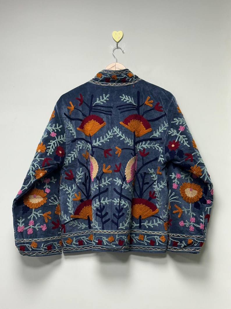 May include: A blue jacket featuring intricate, multi-colored floral embroidery. The design includes large orange, pink, and dark red flower motifs with white leafy stems. The jacket has a high collar and a horizontal patterned border at the hem.