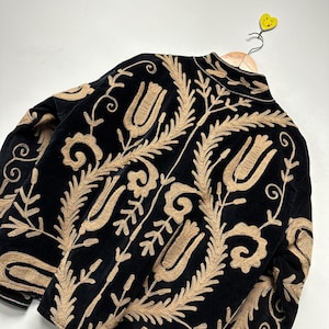 May include: A black velvet jacket featuring intricate tan embroidery in a repeating floral and leaf pattern. The design includes stylised tulip motifs and scrolling vines covering the back and sleeves, with a high collar and a front zip closure.