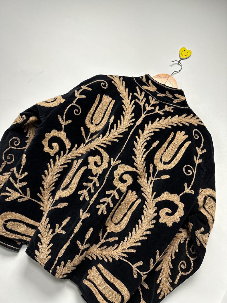 May include: A black velvet jacket featuring intricate tan embroidery in a repeating floral and leaf pattern. The design includes stylised tulip motifs and scrolling vines covering the back and sleeves, with a high collar and a front zip closure.