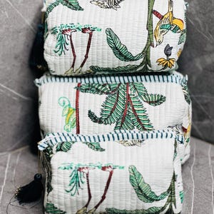 May include: Three white quilted fabric zippered pouches with a green and brown floral print. The pouches are stacked on top of each other. The top pouch has a small tassel.