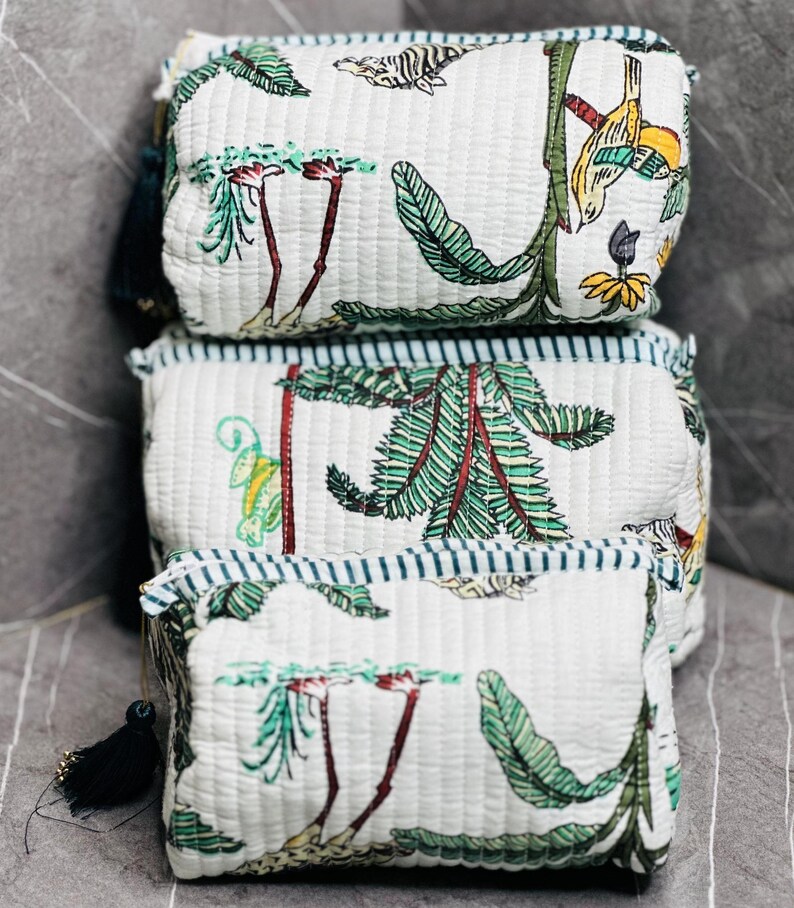 May include: Three white quilted fabric zippered pouches with a green and brown floral print. The pouches are stacked on top of each other. The top pouch has a small tassel.