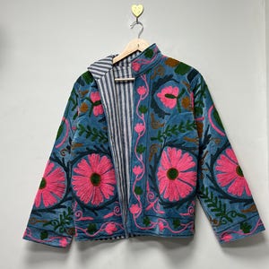 May include: A teal jacket with a floral embroidery design. The jacket features large pink flowers, green leaves, and brown accents. The inside lining is striped in white and blue. The jacket is hanging on a wooden hanger.