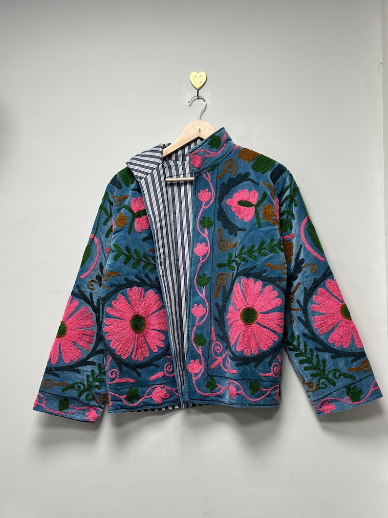 May include: A teal jacket with a floral embroidery design. The jacket features large pink flowers, green leaves, and brown accents. The inside lining is striped in white and blue. The jacket is hanging on a wooden hanger.