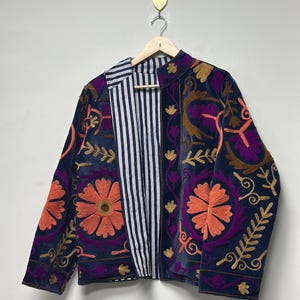 May include: A navy blue jacket featuring vibrant floral embroidery in shades of orange, purple, and gold. The jacket has a high collar and long sleeves, with an interior lining that displays a vertical black and white pinstripe pattern.