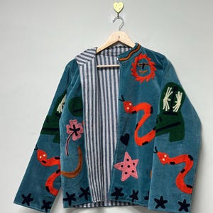 May include: Teal velvet jacket with embroidered orange snakes, stars, and other whimsical designs. The jacket has a striped lining and a collar with orange trim. The jacket is hanging on a wooden hanger.