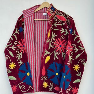 May include: A burgundy velvet jacket featuring vibrant floral embroidery in shades of red, blue, yellow, and green. The interior is lined with a red and white striped fabric. The jacket has long sleeves and an open front, hanging on a wooden hanger.
