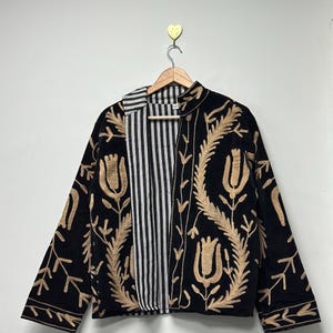 May include: A black jacket featuring tan embroidered floral patterns and a high collar. The interior lining is visible, showcasing a black and white vertical striped fabric. The garment is displayed on a wooden hanger against a plain wall.