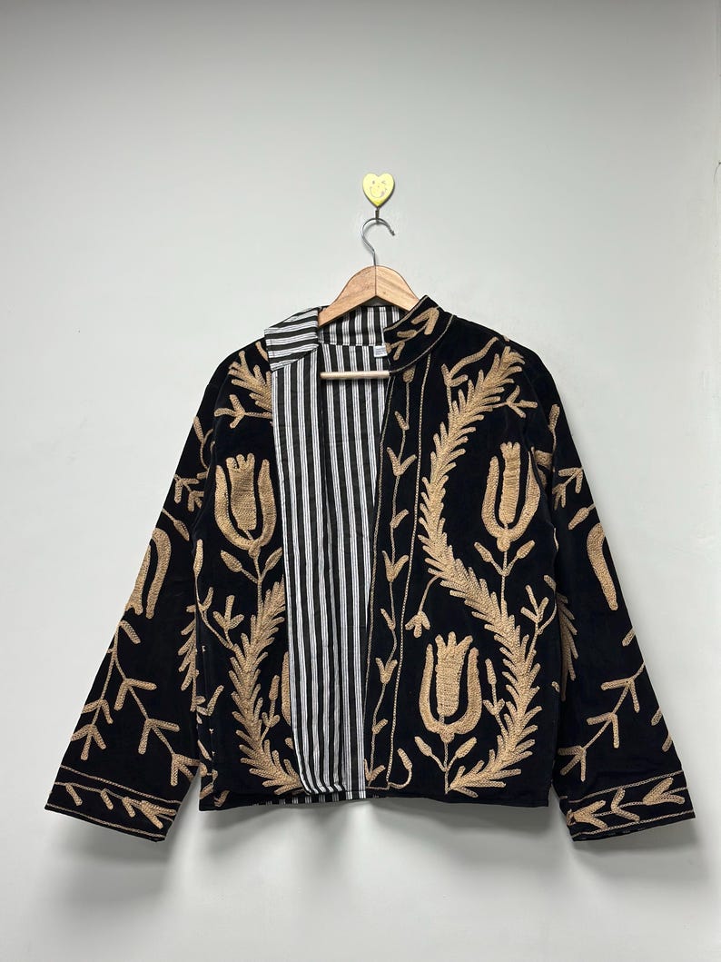 May include: A black jacket featuring tan embroidered floral patterns and a high collar. The interior lining is visible, showcasing a black and white vertical striped fabric. The garment is displayed on a wooden hanger against a plain wall.