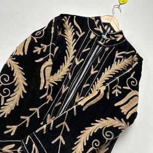 May include: A black velvet jacket featuring intricate gold-coloured embroidery in a leaf and vine pattern. The jacket has a high collar, long sleeves, and a front opening with a metallic clasp closure. The interior lining is white with black stripes.