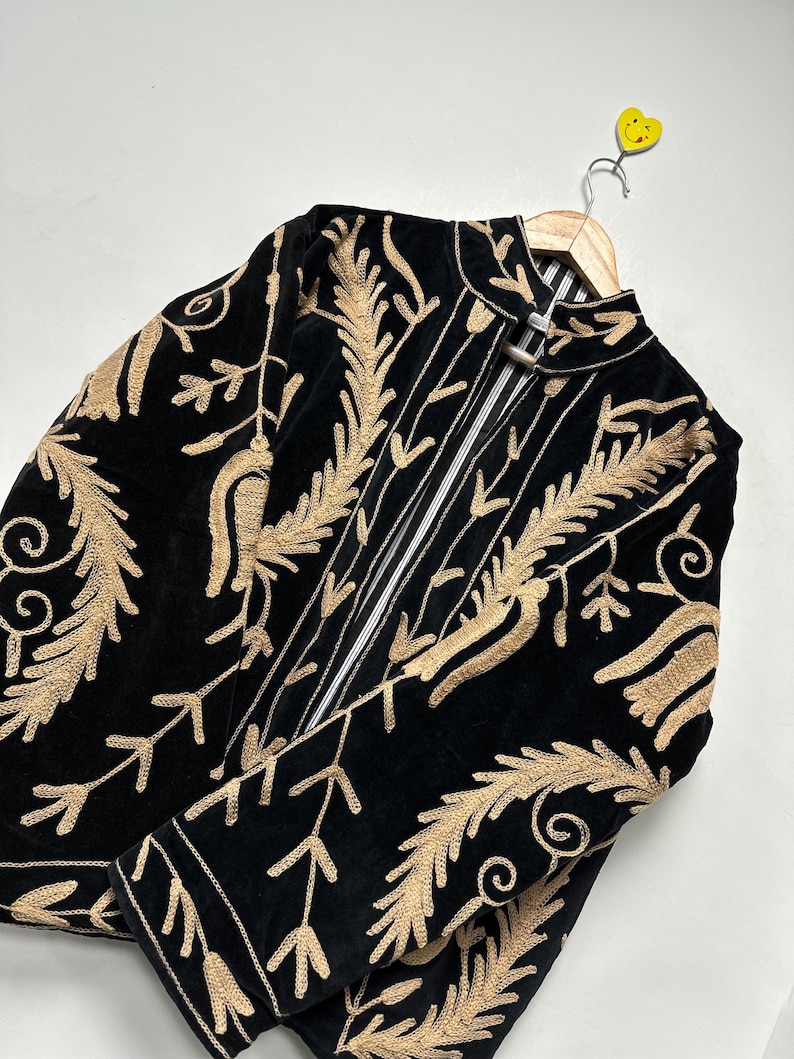 May include: A black velvet jacket featuring intricate gold-coloured embroidery in a leaf and vine pattern. The jacket has a high collar, long sleeves, and a front opening with a metallic clasp closure. The interior lining is white with black stripes.