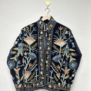 May include: A dark navy blue jacket with intricate floral embroidery in shades of beige, blue, and green. The jacket features a high collar and is hanging on a wooden hanger. The embroidery is detailed and covers the front and sleeves.