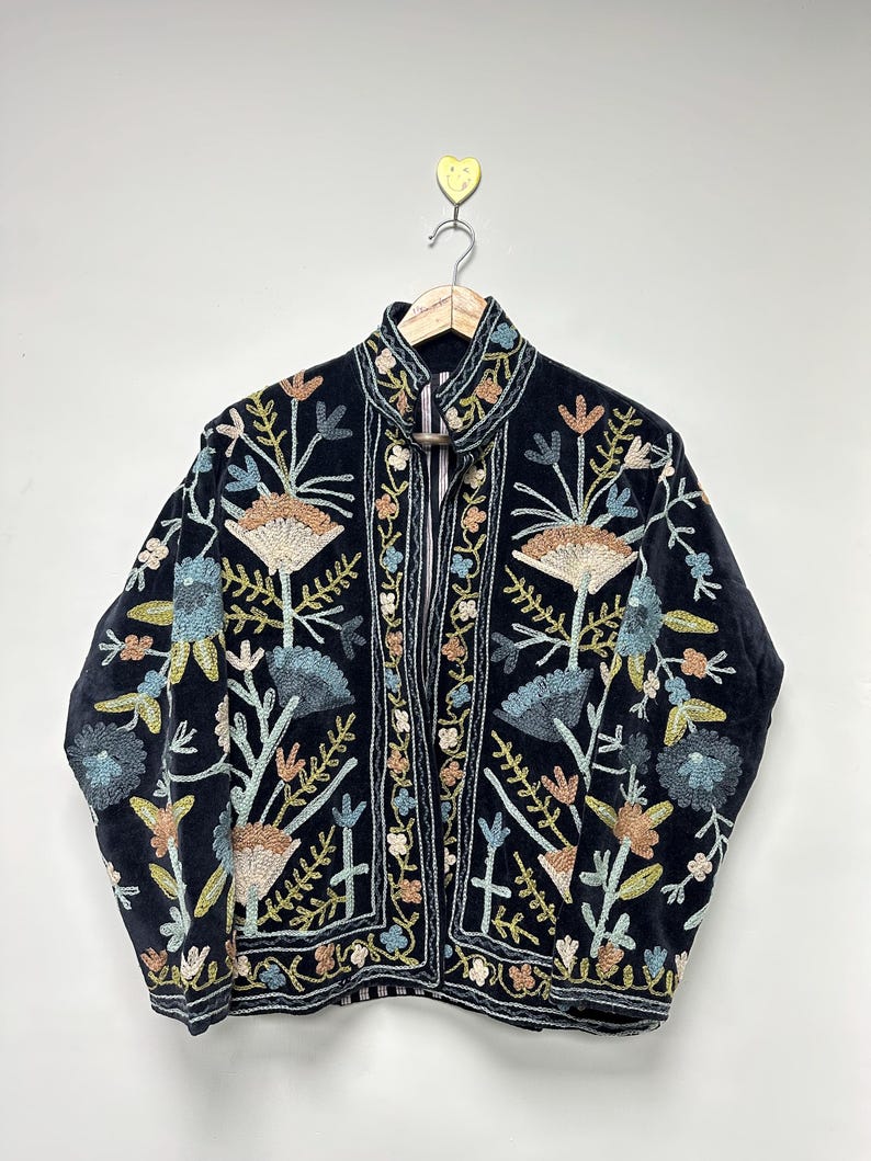 May include: A dark navy blue jacket with intricate floral embroidery in shades of beige, blue, and green. The jacket features a high collar and is hanging on a wooden hanger. The embroidery is detailed and covers the front and sleeves.