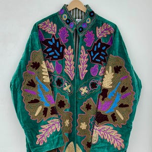 May include: A long-sleeved emerald green velvet jacket featuring intricate, multi-coloured embroidery in shades of purple, blue, gold, and dark brown. The design includes leaf and floral motifs across the front and collar, with a striped lining.