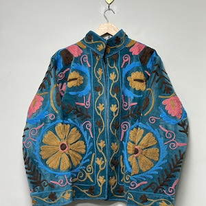 May include: A teal velvet jacket featuring intricate embroidery in shades of mustard yellow, bright pink, and sky blue. The design includes large floral motifs, scrolling vines, and leaf patterns across the front and sleeves, with a high collar.