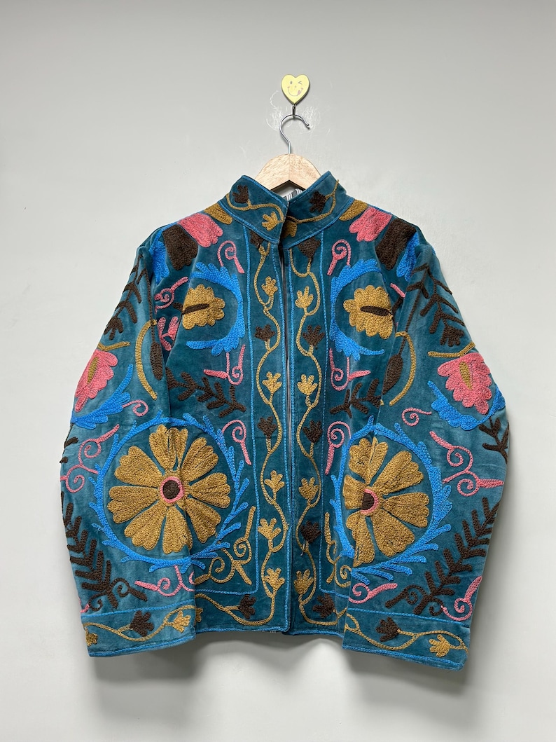 May include: A teal velvet jacket featuring intricate embroidery in shades of mustard yellow, bright pink, and sky blue. The design includes large floral motifs, scrolling vines, and leaf patterns across the front and sleeves, with a high collar.
