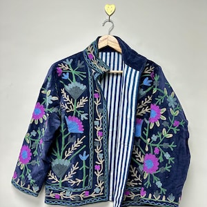 May include: A navy blue velvet jacket with colorful floral embroidery. The jacket features a striped lining and is hanging on a wooden hanger. The embroidery includes pink, blue, and green flowers and vines, creating a bohemian style.