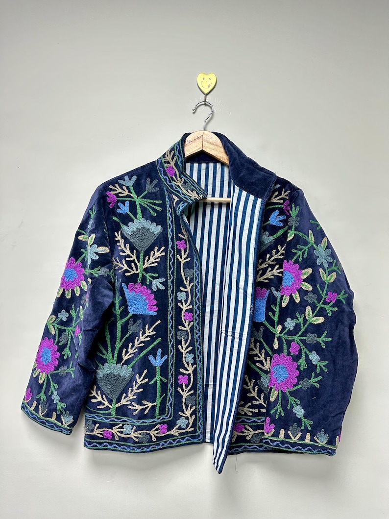May include: A navy blue velvet jacket with colorful floral embroidery. The jacket features a striped lining and is hanging on a wooden hanger. The embroidery includes pink, blue, and green flowers and vines, creating a bohemian style.