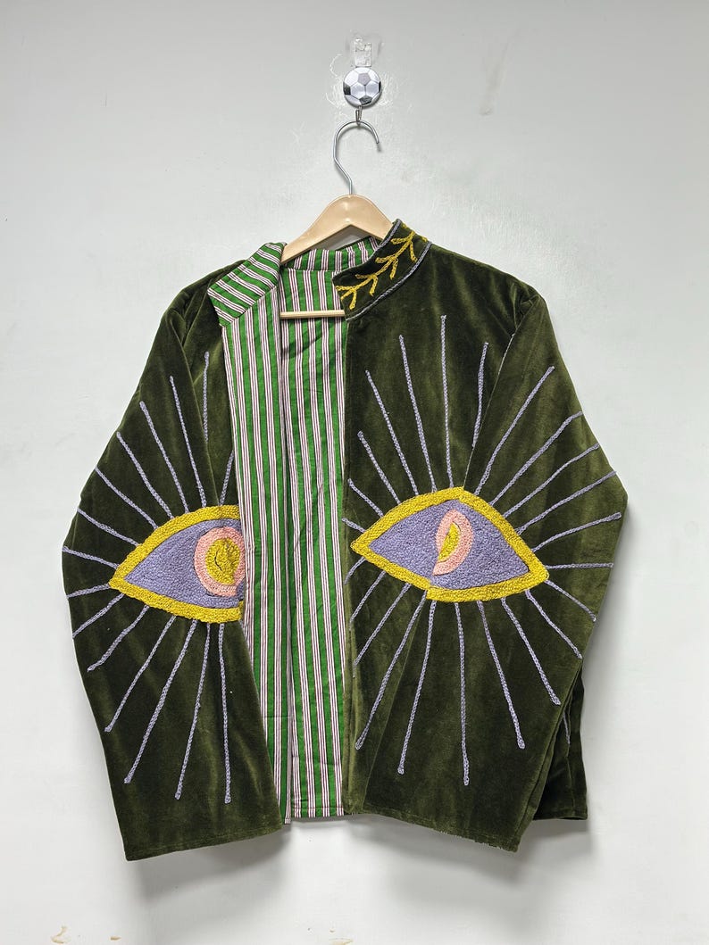 K&ouml;nnte beinhalten: Eine olivgr&uuml;ne Samtjacke mit gestickten Augenmotiven auf den Vorderteilen. Die Augen sind in lavendelfarbenem, rosa und gelbem Garn mit strahlenden silbernen Linien ausgef&uuml;hrt. Das Innenfutter zeigt ein gr&uuml;n-wei&szlig;es vertikales Streifenmuster.