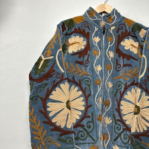 May include: A light blue velvet jacket featuring intricate embroidery in cream, dark brown, and olive green. The design showcases large floral motifs, swirling vines, and leaf patterns across the front panels, sleeves, and high collar.
