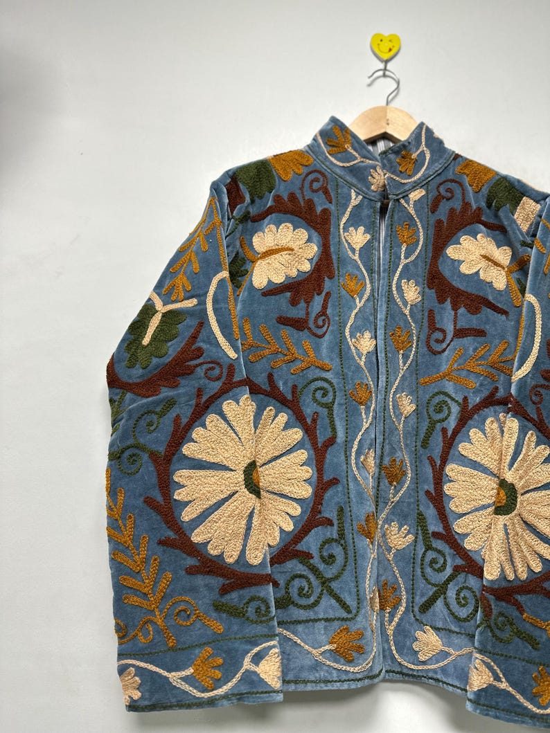 May include: A light blue velvet jacket featuring intricate embroidery in cream, dark brown, and olive green. The design showcases large floral motifs, swirling vines, and leaf patterns across the front panels, sleeves, and high collar.