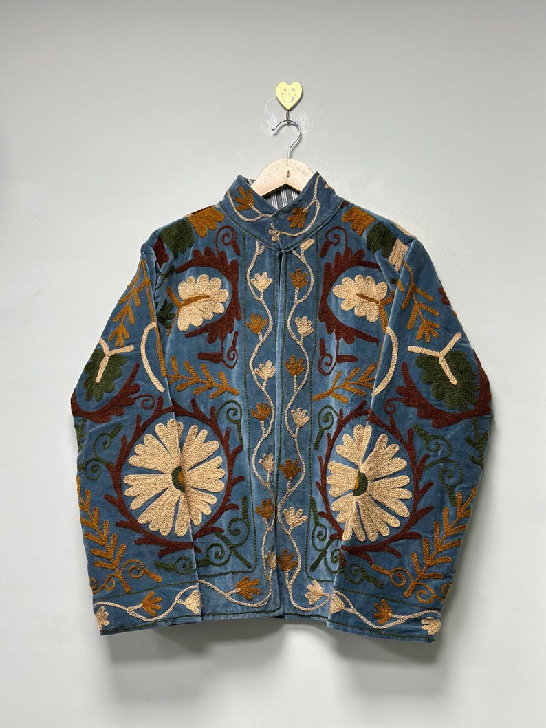 May include: A blue velvet jacket featuring intricate embroidery in cream, dark brown, and olive green. The design includes large floral motifs, scrolling vines, and leaf patterns across the front and sleeves, with a high collar and long sleeves.