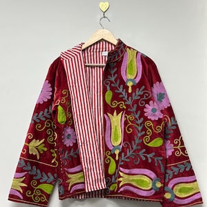 May include: A red velvet jacket with floral embroidery in green, pink, and yellow. The jacket has a striped red and white lining and a high collar. The jacket is hanging on a wooden hanger.