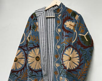 May include: A blue jacket featuring intricate floral embroidery in shades of tan, brown, and green. The garment has a high collar and a front opening that reveals a black and white vertical striped lining. It is displayed on a wooden hanger.
