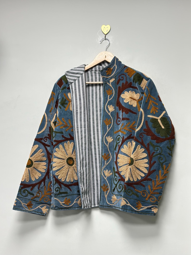 May include: A blue jacket featuring intricate floral embroidery in shades of tan, brown, and green. The garment has a high collar and a front opening that reveals a black and white vertical striped lining. It is displayed on a wooden hanger.