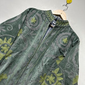 May include: A green velvet jacket with intricate embroidered floral patterns in gray and lime green. The jacket features a mandarin collar and a striped lining. It is displayed on a wooden hanger.