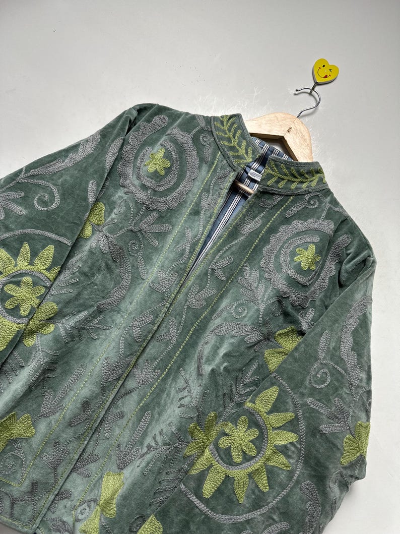 May include: A green velvet jacket with intricate embroidered floral patterns in gray and lime green. The jacket features a mandarin collar and a striped lining. It is displayed on a wooden hanger.