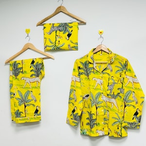 Puede incluir: Conjunto de pijama amarillo con estampado de animales de la selva, incluyendo cebras, leopardos y tucanes. El conjunto incluye una camisa con botones, pantalones y una m&aacute;scara para los ojos, todo colgado en perchas de madera.