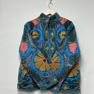 May include: A teal jacket featuring intricate embroidery in shades of mustard yellow, bright pink, and dark brown. The garment has a high collar, long sleeves, and a front zipper closure, with floral and vine patterns covering the entire surface.