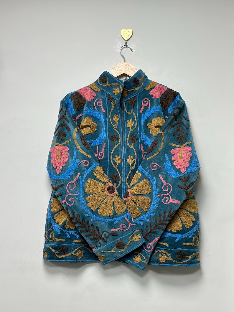 May include: A teal jacket featuring intricate embroidery in shades of mustard yellow, bright pink, and dark brown. The garment has a high collar, long sleeves, and a front zipper closure, with floral and vine patterns covering the entire surface.