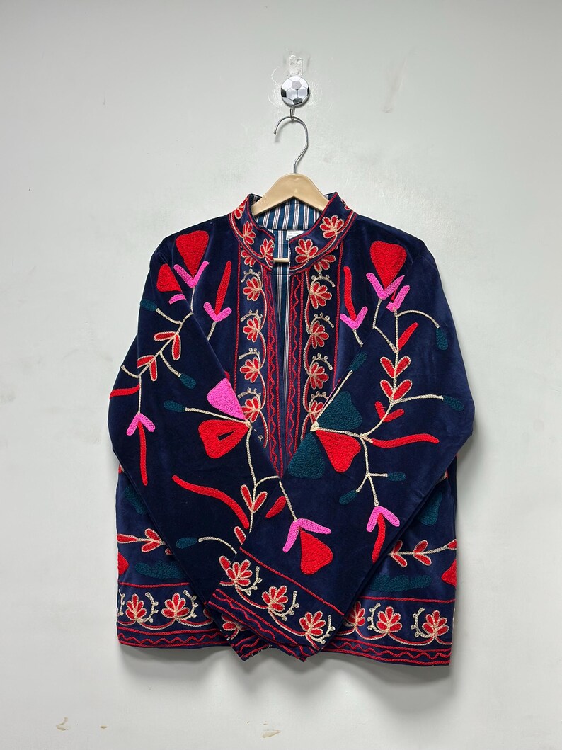 Op de afbeelding: Een donkerblauw fluwelen jasje met levendige bloemenborduursels in rood, roze en petrol. Het jasje heeft een mandarijnkraag, lange mouwen en een gestreepte voering, met symmetrische botanische patronen op de voorpanden van het kledingstuk.