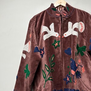 May include: A brown velvet jacket with embroidered white birds, blue hearts, green leaves, and orange stars. The jacket has a high collar and a zipper closure. The jacket is hanging on a wooden hanger.