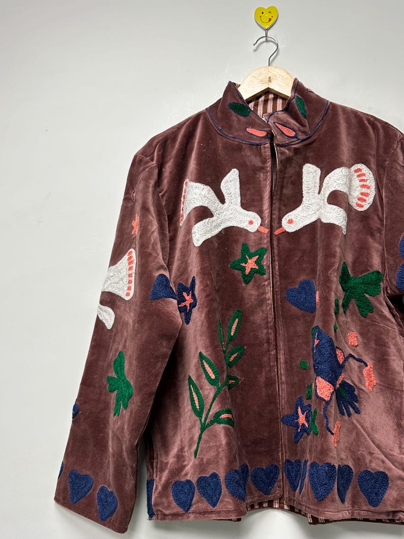 May include: A brown velvet jacket with embroidered white birds, blue hearts, green leaves, and orange stars. The jacket has a high collar and a zipper closure. The jacket is hanging on a wooden hanger.