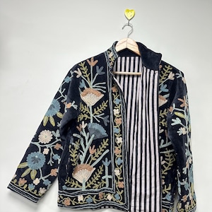 May include: A black velvet jacket with colorful floral embroidery. The jacket features a striped interior and is hanging on a wooden hanger. The embroidery includes shades of blue, peach, and green.