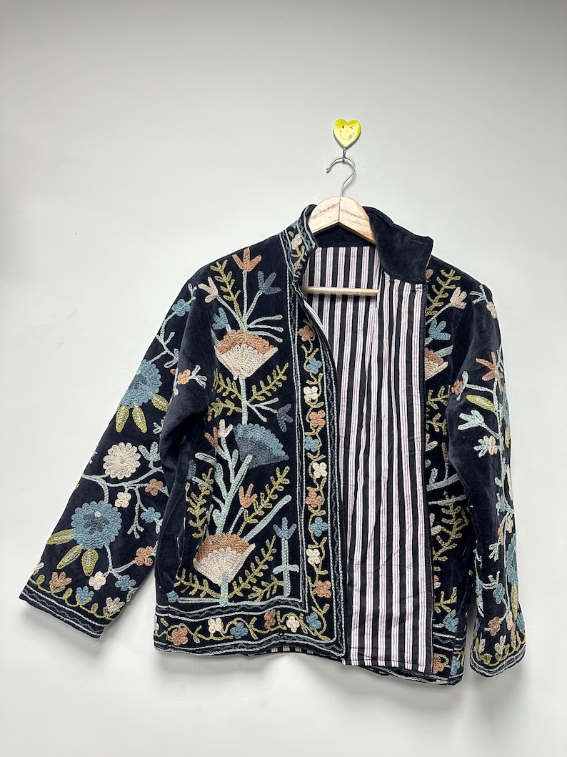 May include: A black velvet jacket with colorful floral embroidery. The jacket features a striped interior and is hanging on a wooden hanger. The embroidery includes shades of blue, peach, and green.