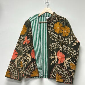 May include: A dark green velvet jacket with floral embroidery in orange, gold, and beige. The jacket has a striped green and white lining and is hanging on a wooden hanger.