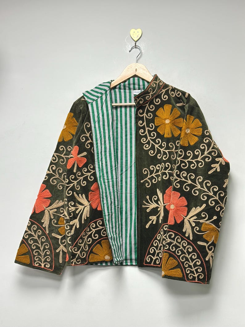 May include: A dark green velvet jacket with floral embroidery in orange, gold, and beige. The jacket has a striped green and white lining and is hanging on a wooden hanger.