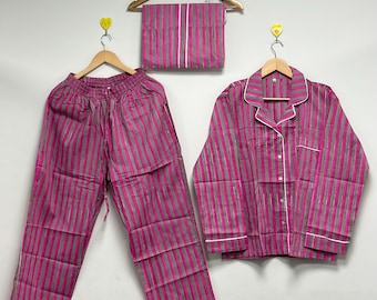 Puede incluir: Un conjunto de pijama a rayas rosas y grises, que incluye pantalones, una camisa con botones y una sábana doblada. El conjunto presenta ribetes blancos y se exhibe en perchas de madera contra una pared blanca.