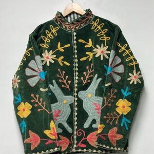 May include: A dark green velvet jacket featuring a high collar and intricate embroidery. The design includes two light blue rabbits facing each other, surrounded by colorful floral and leaf patterns in yellow, blue, and pink thread.