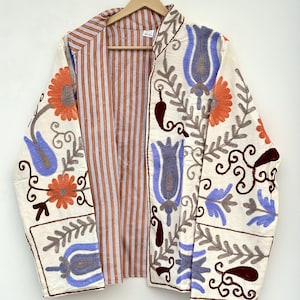 May include: A cream-coloured jacket featuring vibrant floral and botanical embroidery in shades of orange, purple, and dark red. The interior lining displays a vertical brown and white striped pattern. The garment is displayed on a wooden hanger.